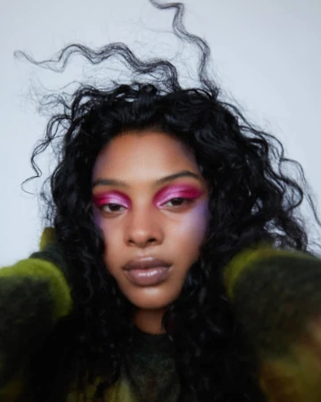 Close-up portrait of a woman with long dark curly hair wearing vibrant pink eyeshadow against a white background by Anairam