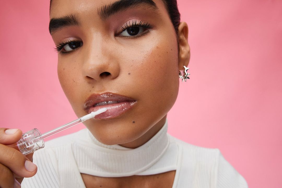 Close-up of a young adult woman with pink eyeshadow applying clear lip gloss in a photo studio setting against a pink background.