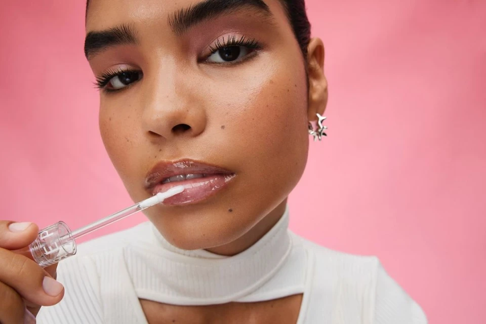 Close-up of a young adult woman with pink eyeshadow applying clear lip gloss in a photo studio setting against a pink background.
