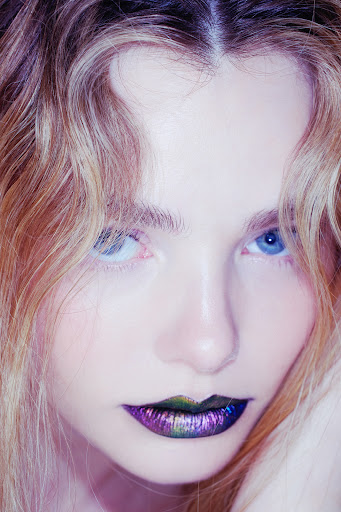 Close-up portrait of a woman with vibrant purple and metallic lips, blue eyes, and wavy blonde-highlighted hair.