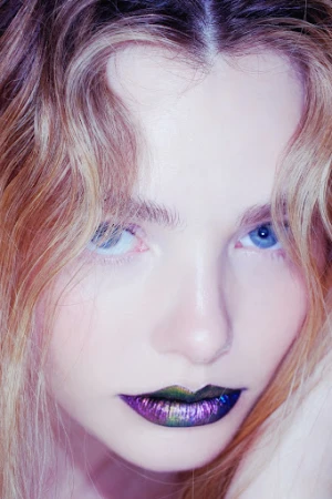 Close-up portrait of a woman with vibrant purple and metallic lips, blue eyes, and wavy blonde-highlighted hair.
