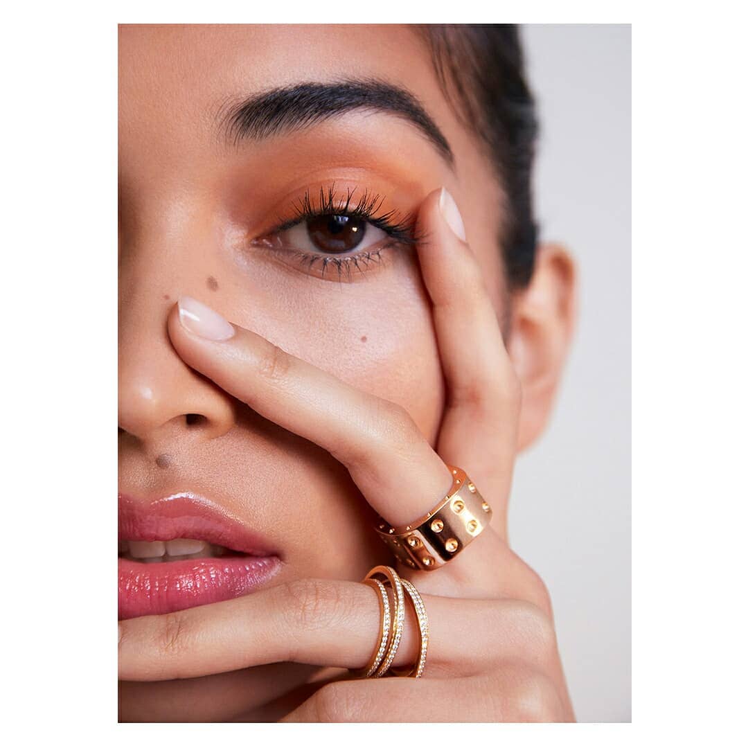 Close-up of a woman with tan eye shadow and glossy lips, covering one eye with her fingers and wearing gold rings on her hand.