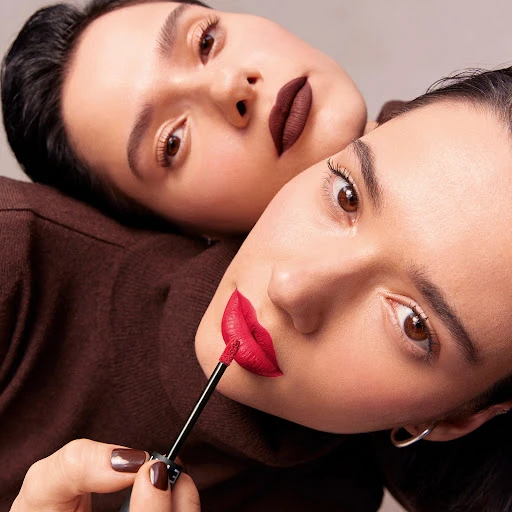 Two women pose closely together, one applying red lipstick with a brush. A beauty shot for makeup or cosmetics advertising, indoors.