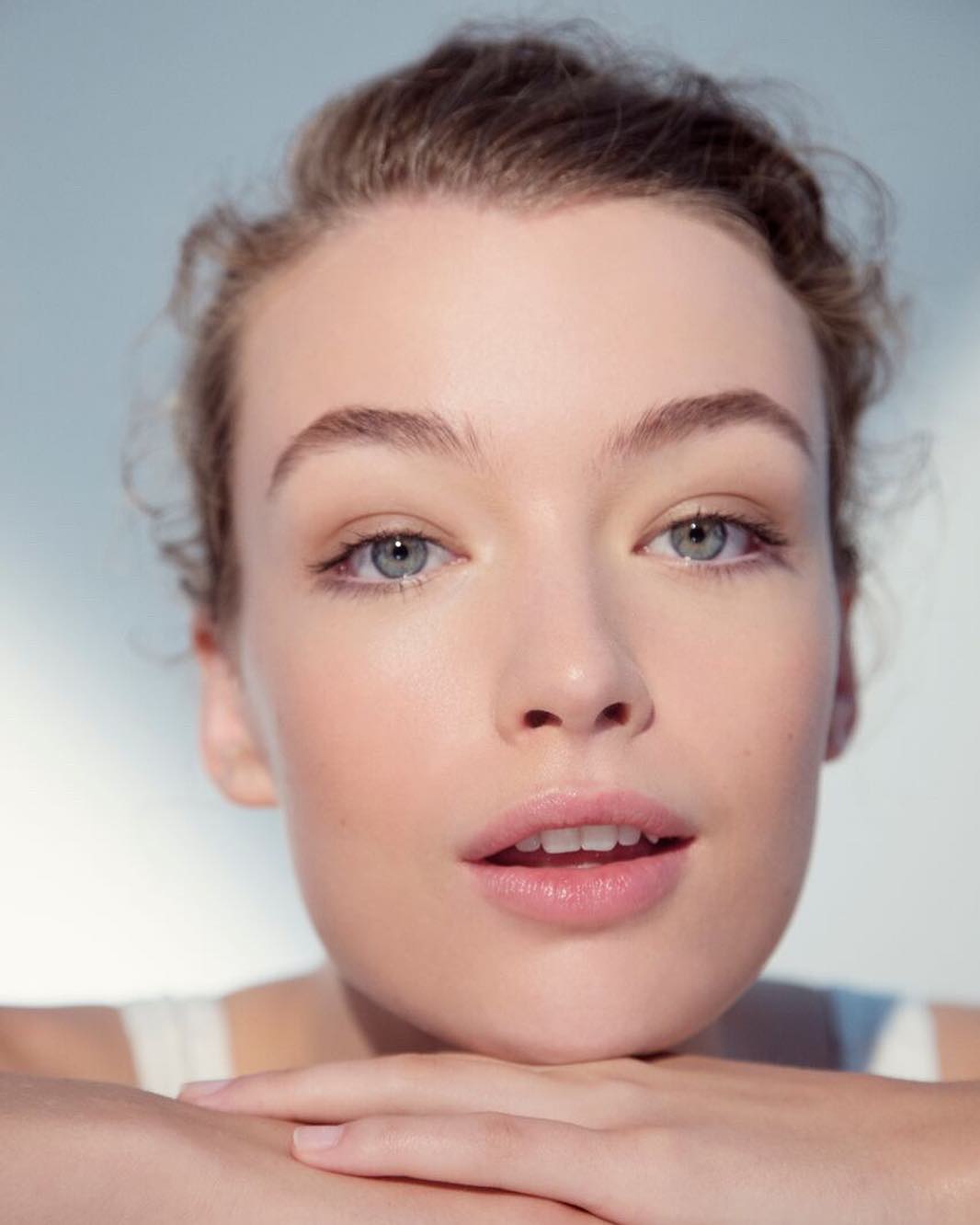 Close-up portrait of a fair-skinned young woman with light eyes and her hands clasped beneath her chin, against a pale blue background.
