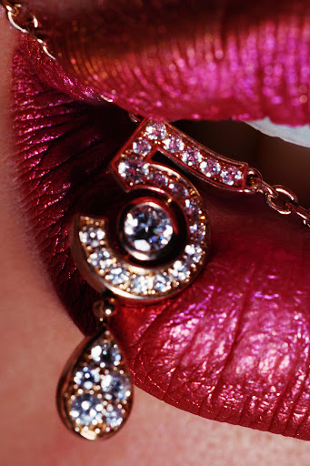 Close-up of lips with shimmering fuchsia lipstick and a gold necklace featuring a jeweled number five pendant with a teardrop gem.