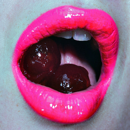 Close-up of a woman's mouth with bright pink lipstick holding two dark cherries between her teeth, creating a bold and sensual image.
