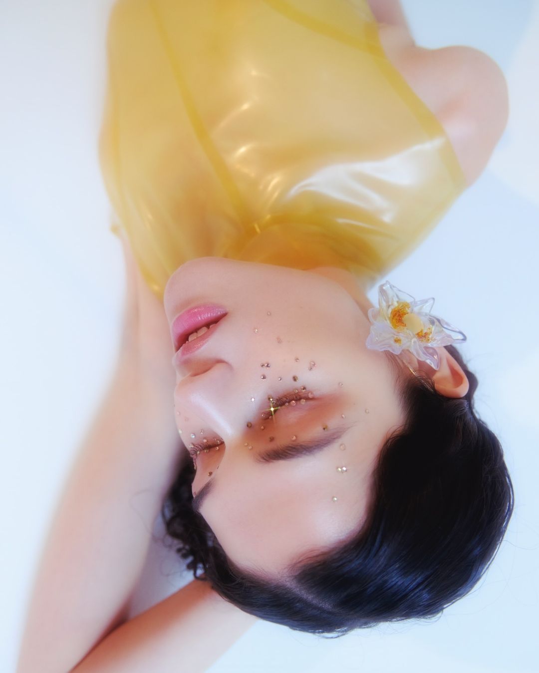 Artistic portrait of a woman with glitter makeup and a clear yellow top, lying in a light blue setting with an ornate floral earring.