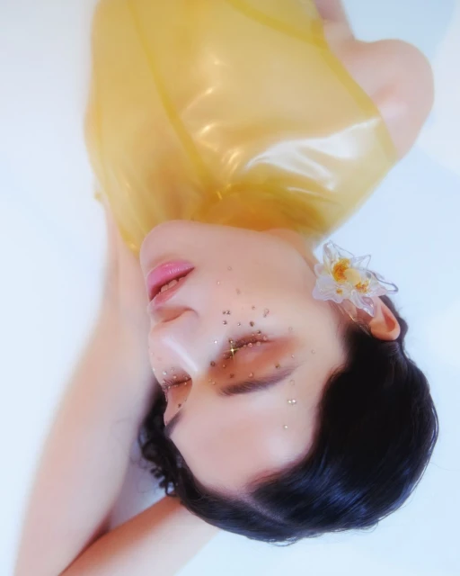 Artistic portrait of a woman with glitter makeup and a clear yellow top, lying in a light blue setting with an ornate floral earring.