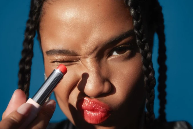 Close-up of a woman with braided hair applying red lipstick, winking and posing against a vibrant blue backdrop.