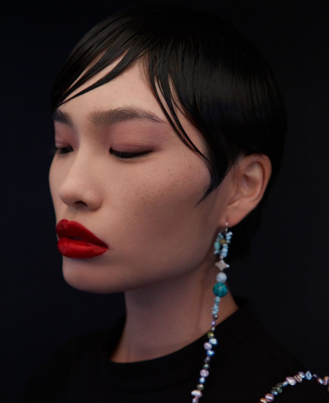 Close-up shot of a woman with red lipstick and short black hair, wearing a beaded earring against a dark background.