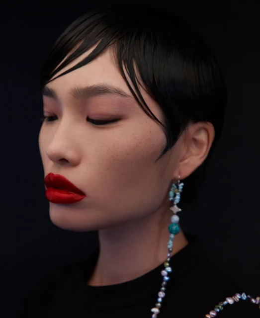 Close-up shot of a woman with red lipstick and short black hair, wearing a beaded earring against a dark background.