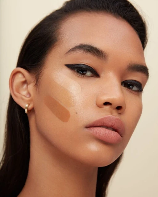 Close-up of a woman with tan skin wearing makeup, including black eyeliner and foundation swatches in varying shades on her cheek.