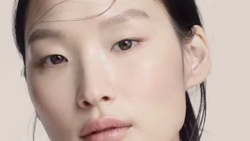 Close-up portrait of an Asian woman with light makeup and slightly wet hair, set against a neutral, monochromatic background.
