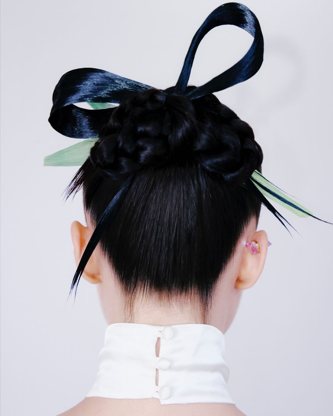 Back view of a woman with braided black hair adorned with dark blue and light green ribbons, wearing a white high-necked top against a light background.