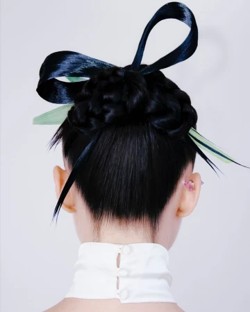 Back view of a woman with braided black hair adorned with dark blue and light green ribbons, wearing a white high-necked top against a light background.