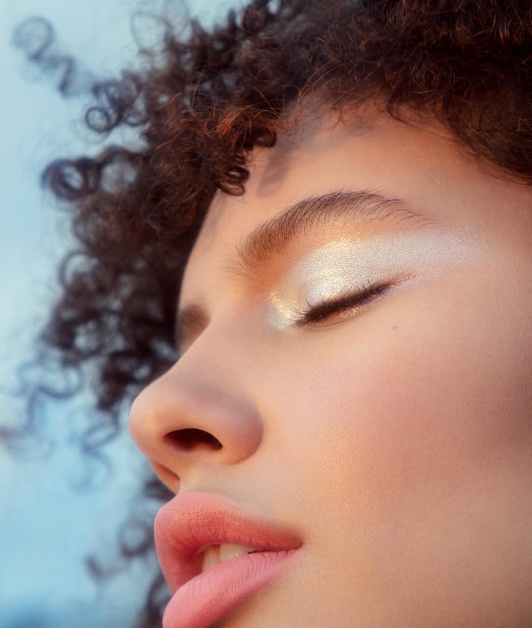Close-up of a woman's face with makeup. Golden eyeshadow and pink lips. Soft lighting accentuates her features against a blurred sky backdrop.