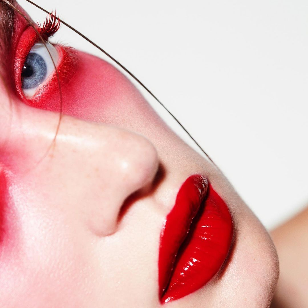 Close-up beauty portrait of a model with vibrant red eye makeup, glossy red lips, and a single strand of hair across the face.