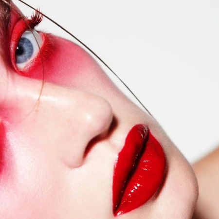Close-up beauty portrait of a model with vibrant red eye makeup, glossy red lips, and a single strand of hair across the face.
