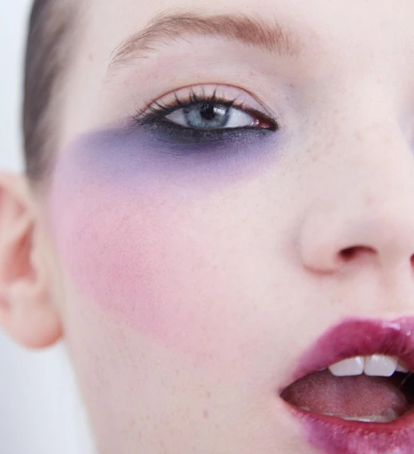 Close-up studio shot of a fair-skinned adult female model with purple eye shadow and dark pink lipstick against a white background.
