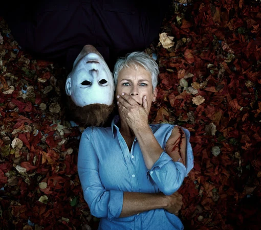 Jamie Lee Curtis with Halloween killer mask on ground surrounded by fall leaves; she covers her mouth with hand looking shocked.