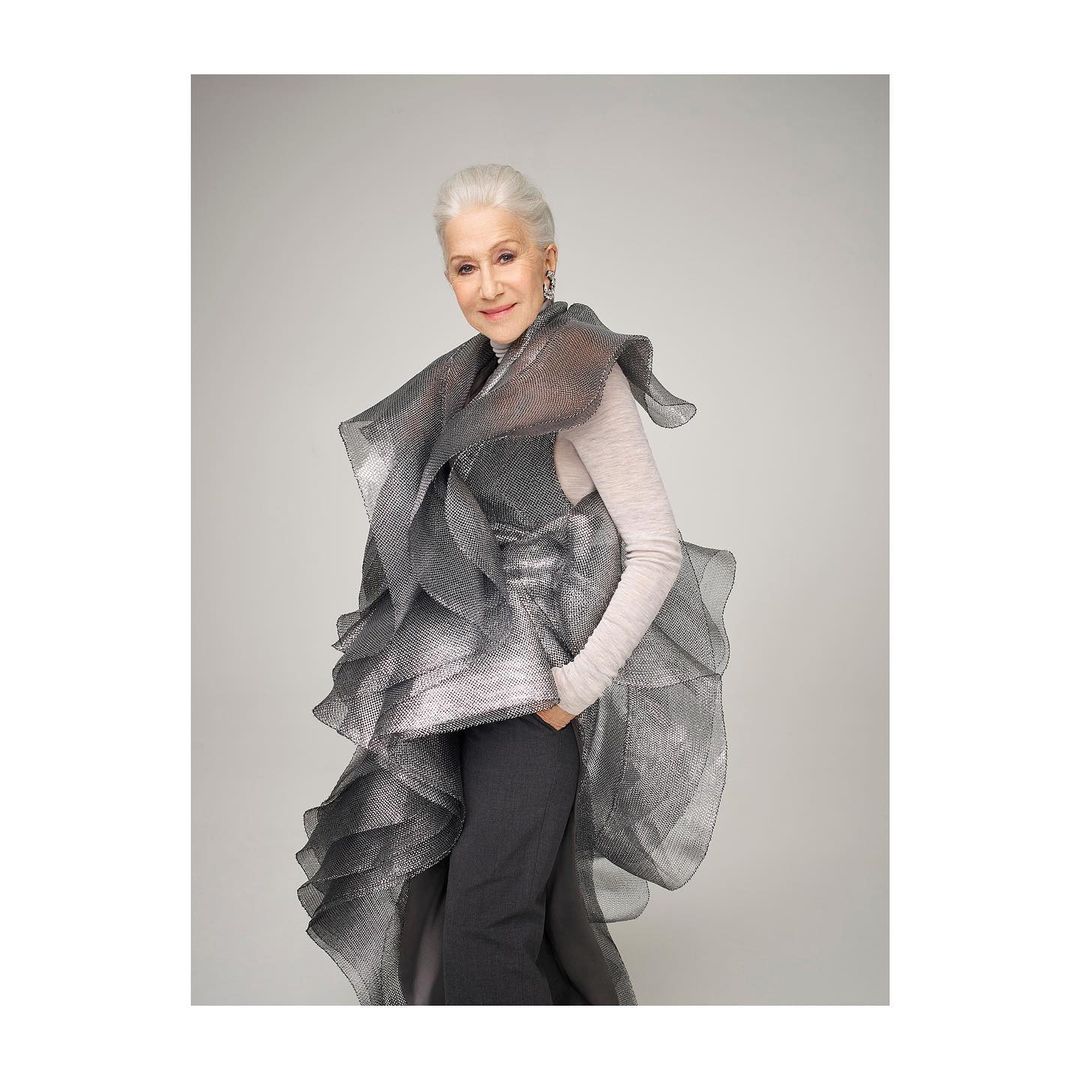 Older woman with short grey hair posing in a dramatic ruffled grey gown against a neutral background in a fashion forward portrait.
