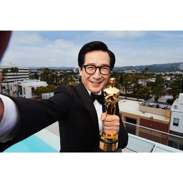 Man in tuxedo takes selfie with Oscar statue on rooftop with urban background, smiling broadly; awards, celebrity, achievement.