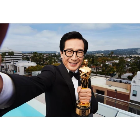 Man in tuxedo takes selfie with Oscar statue on rooftop with urban background, smiling broadly; awards, celebrity, achievement.