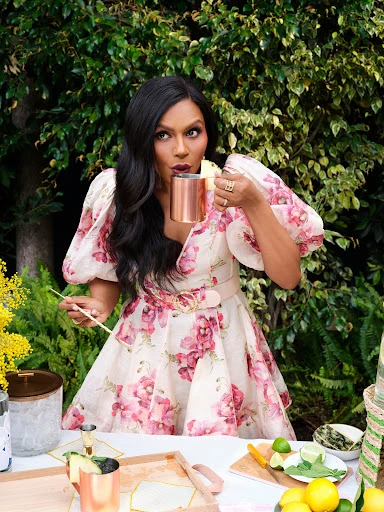 A woman in a floral dress sips from a copper mug outdoors amid greenery and citrus fruit, creating a vibrant, summery scene.
