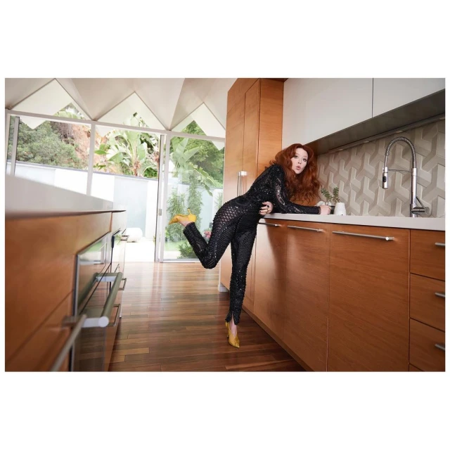 A redhead woman in a black sequin catsuit and yellow heels strikes a pose inside a modern kitchen with natural lighting.