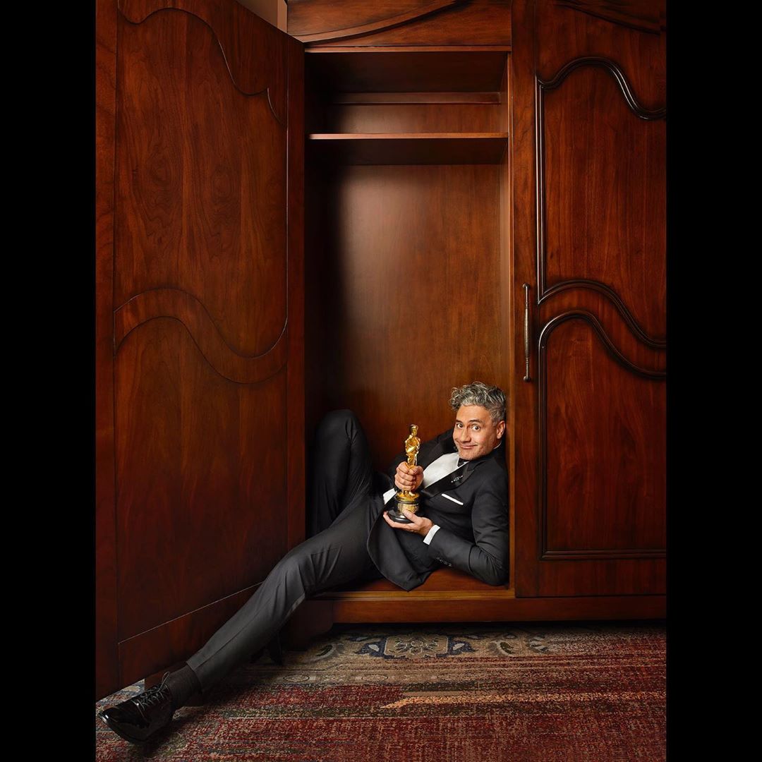 Adult male in a suit lying inside of a wooden wardrobe holding a golden statuette trophy in an indoor environment with a rug on the floor.