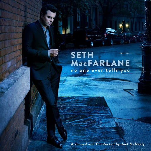 Seth MacFarlane in a dark suit leans against a brick wall while smoking a cigarette, under streetlight on a city street at night.