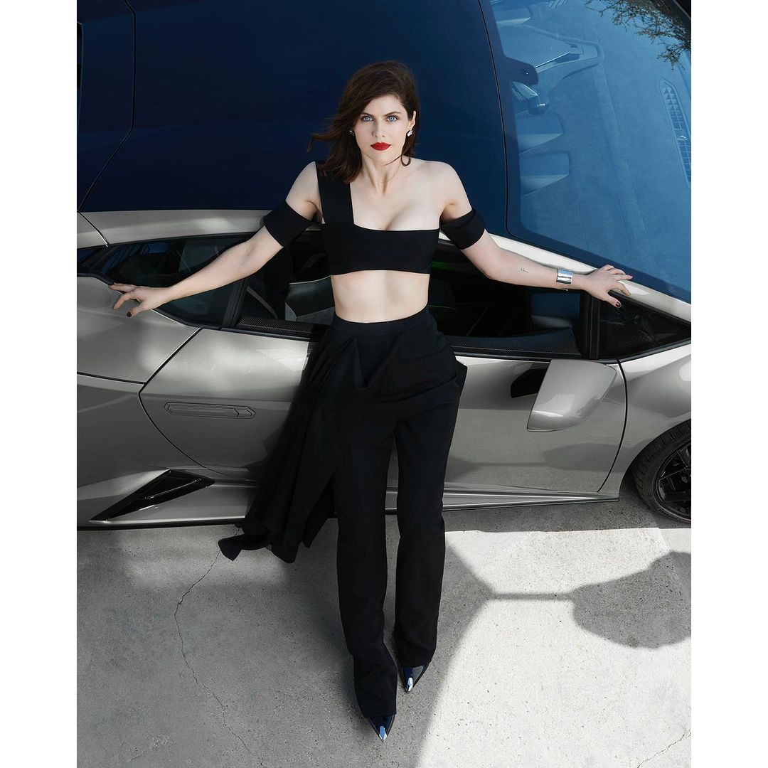 Alexandra Daddario poses in black outfit alongside a silver Lamborghini against a blurred backdrop, showcasing fashion and automotive elements.