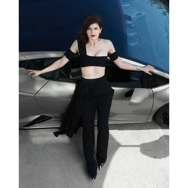 Alexandra Daddario poses in black outfit alongside a silver Lamborghini against a blurred backdrop, showcasing fashion and automotive elements.