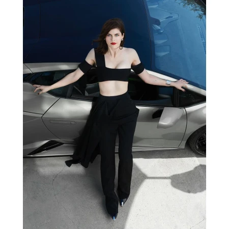 Alexandra Daddario poses in black outfit alongside a silver Lamborghini against a blurred backdrop, showcasing fashion and automotive elements.