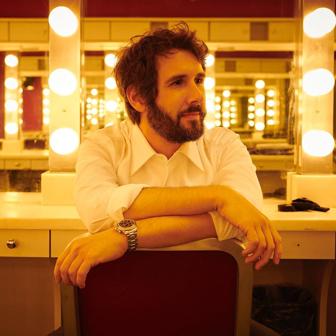 Portrait of a man with a beard and wrist watch sitting in front of a vanity mirror with lights in an indoor setting.