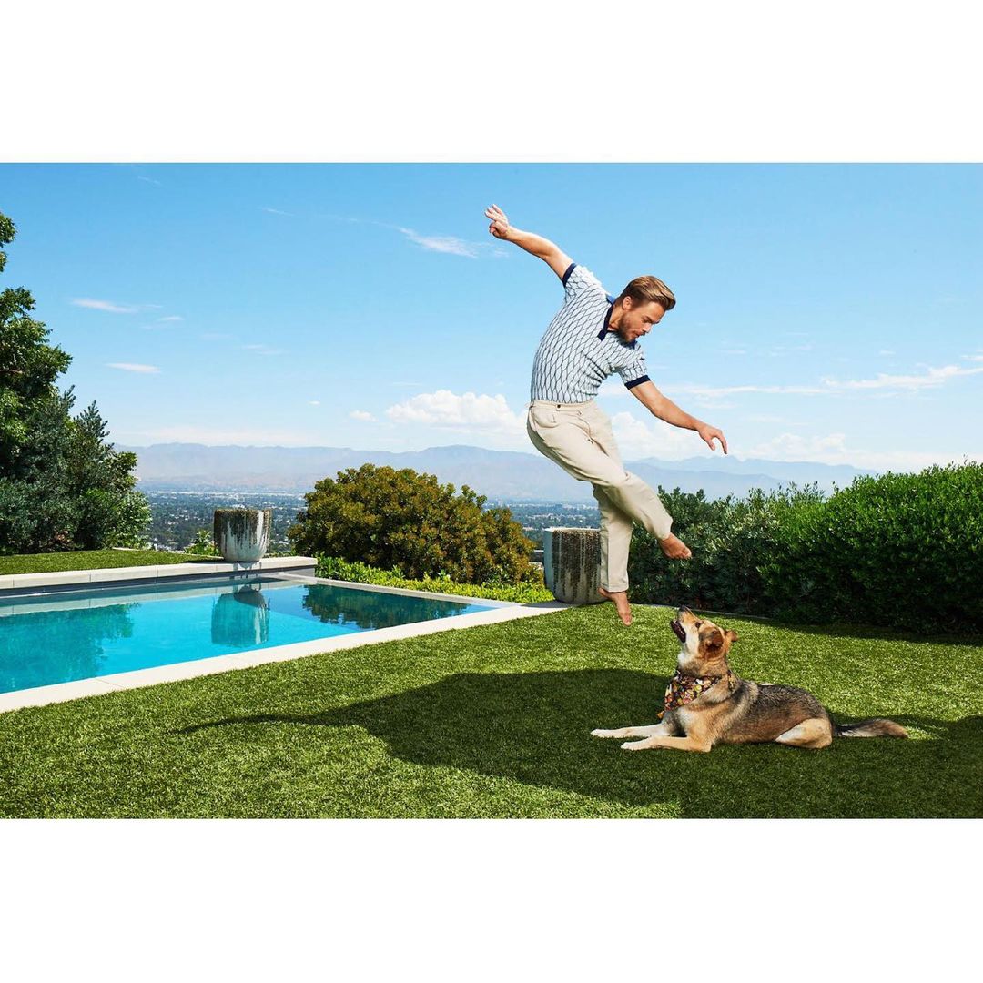 An adult man balances on one leg near a swimming pool, with a dog looking up at him, under a bright blue sky in a sunny outdoor environment.