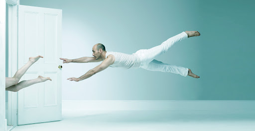 Bald man in white clothing mid-air with outstretched arms, another person's legs visible through an open door, set against a light blue background.