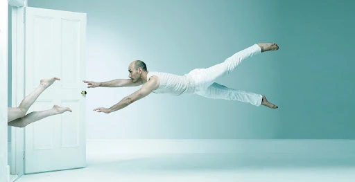 Bald man in white clothing mid-air with outstretched arms, another person's legs visible through an open door, set against a light blue background.