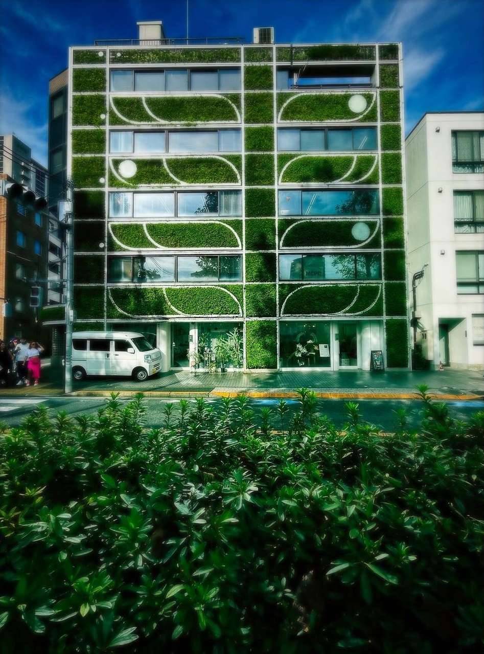 Modern building facade covered in lush green plants with white trim, blue sky overhead and parked van on a city street.