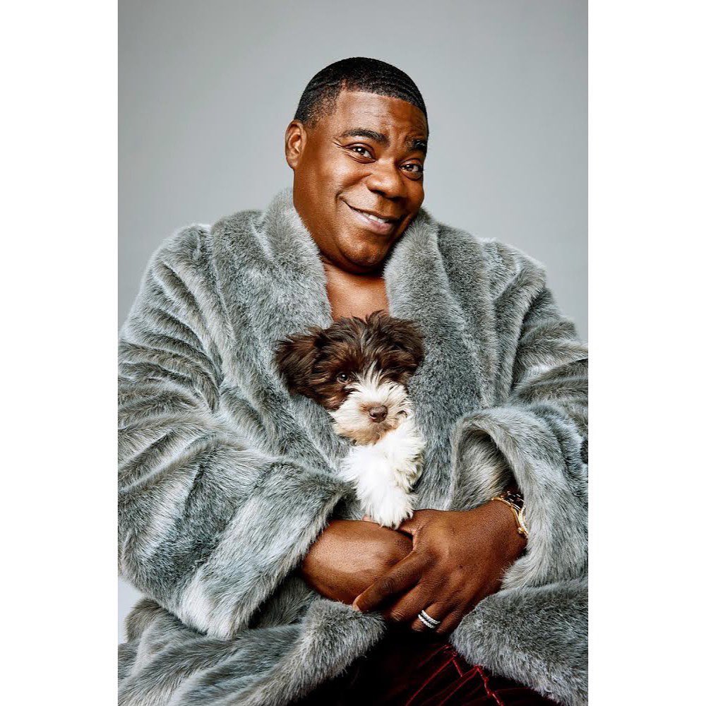 Portrait of an adult male smiling and holding a small brown-and-white dog while wearing a large grey fur coat against a grey backdrop.