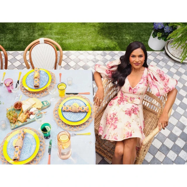 Mindy Kaling poses in floral dress next to outdoor table setting with bright colors, plates, and beverages on a sunny day.