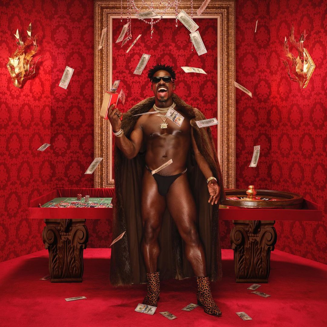 A muscular shirtless adult male in underwear stands in front of a casino table with money falling in a red room with gold accents.