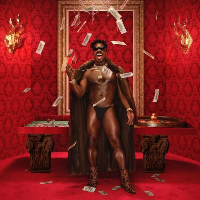 A muscular shirtless adult male in underwear stands in front of a casino table with money falling in a red room with gold accents.