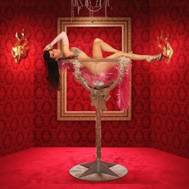 A woman poses in a large champagne glass on a red surface with a red patterned wall and gold decor for a glamourous photo shoot.