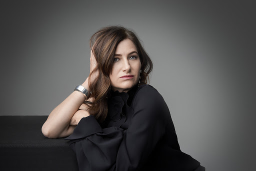 Portrait of actress Kathryn Hahn resting her head on her hand, wearing a dark blouse and a silver cuff bracelet against a grey backdrop.