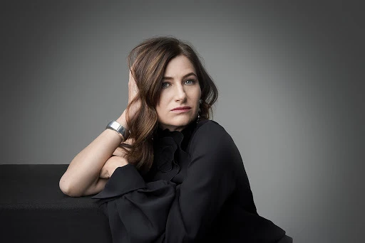 Portrait of actress Kathryn Hahn resting her head on her hand, wearing a dark blouse and a silver cuff bracelet against a grey backdrop.