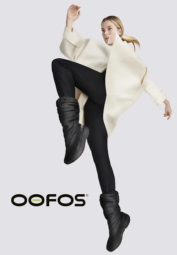 Athletic woman wearing black leggings, black puffer boots and a cream coat is jumping in a studio setting to advertise OOFOS boots.