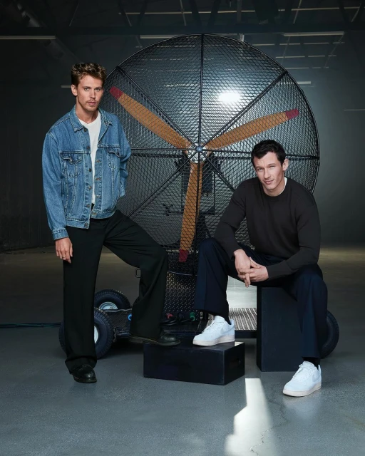 Two adult men pose indoors in a studio setting with a large vintage fan prop. One man stands in a denim jacket, the other sits on a cube.