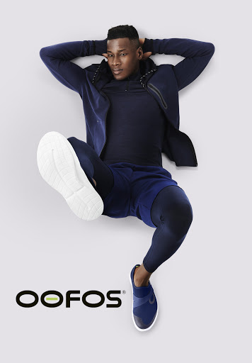 Athletic African American man in navy activewear lying on a gray surface with OOFOS logo. Fitness fashion product shot.
