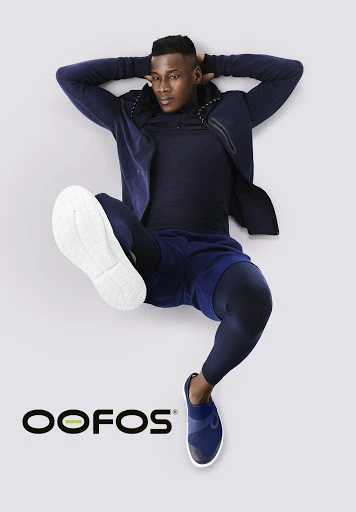 Athletic African American man in navy activewear lying on a gray surface with OOFOS logo. Fitness fashion product shot.